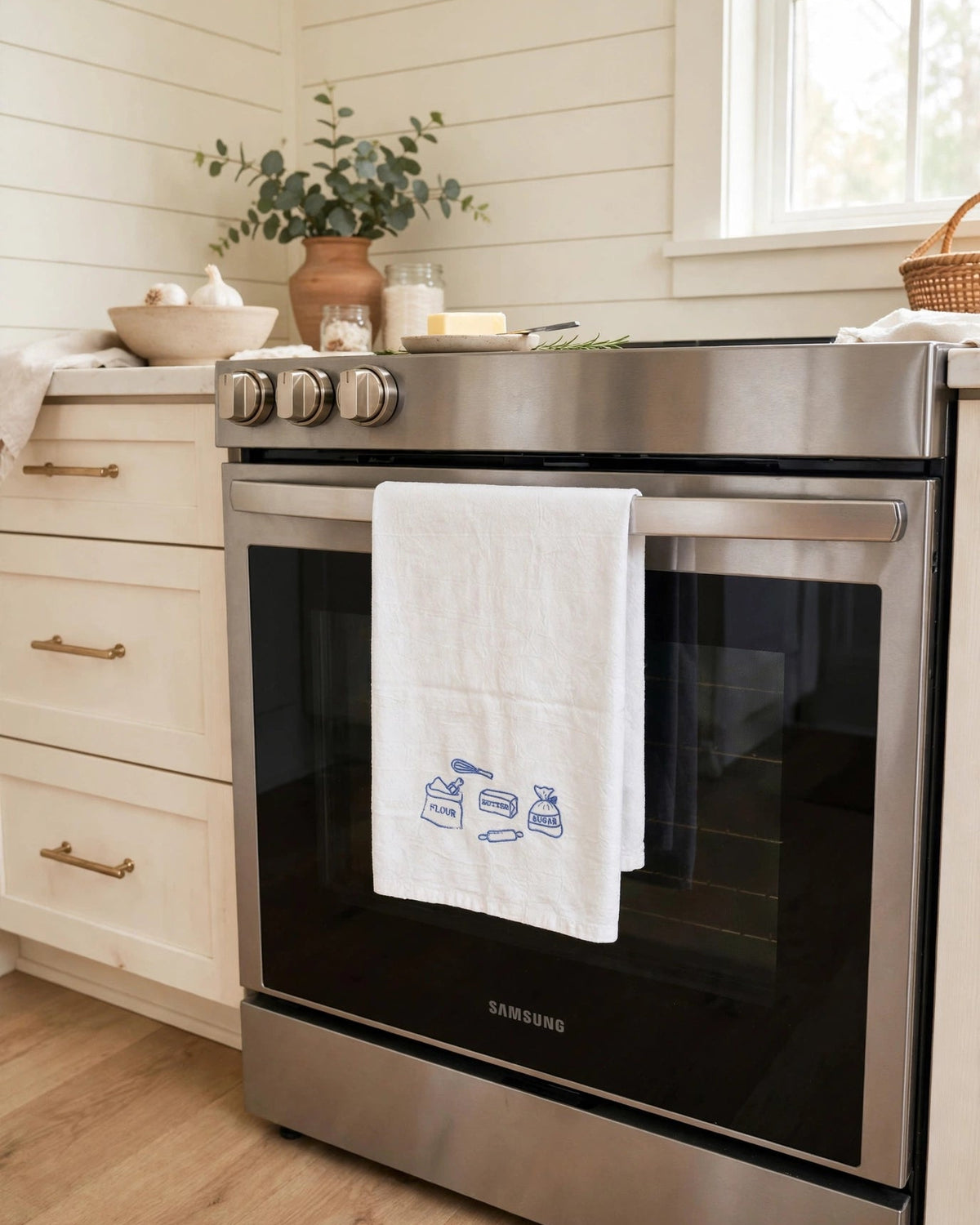 White embroidered tea towel with baking design in blue thread hanging on a stainless oven handle in a bright kitchen