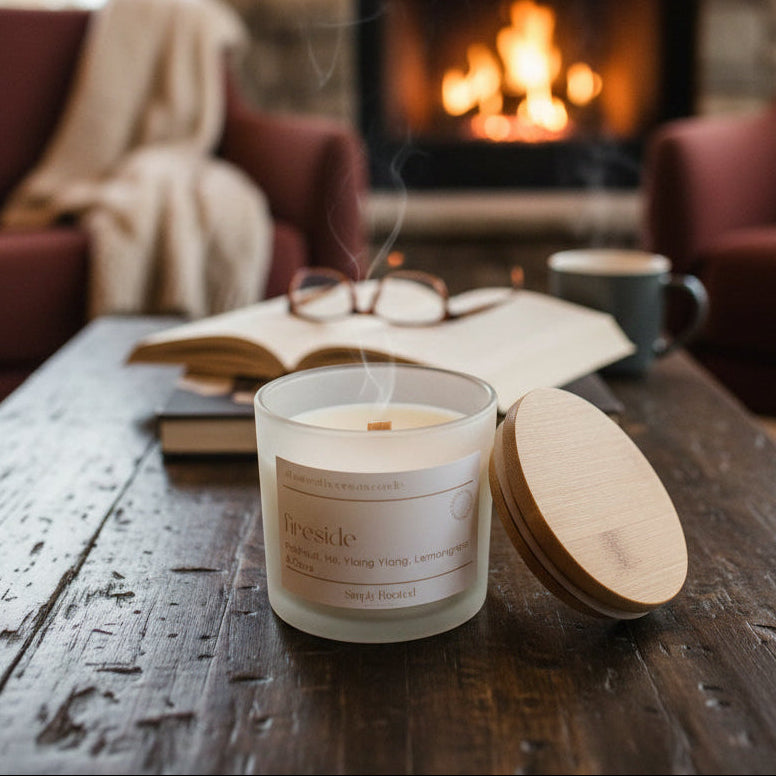 A lit scented candle in a frosted glass jar sits on a rustic wooden table in a cozy living room setting, with a burning fireplace, an open book, glasses, and a mug in the background. The candle label reads 'feeside' and 'Scented Wax Co.