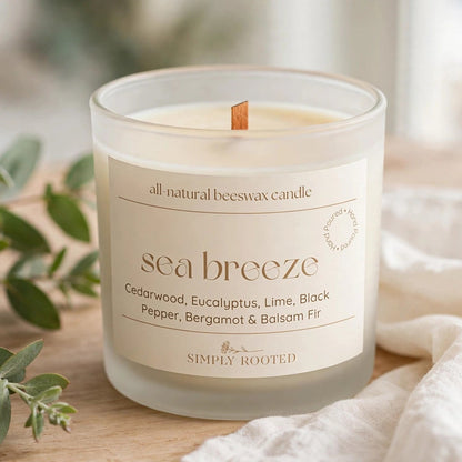 A glass jar candle labeled 'sea breeze' by Simply Rooted, placed on a wooden surface with eucalyptus leaves and a white fabric napkin in a softly lit setting.