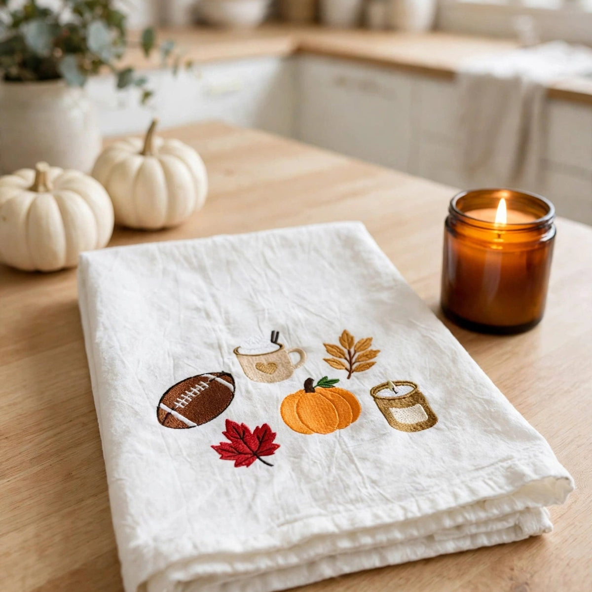 A folded white linen kitchen towel with autumn-themed embroidery sits on a wooden table in a warm kitchen setting. The embroidery features a football, a mug, a pumpkin, a leaf, a candle jar, and a red maple leaf.