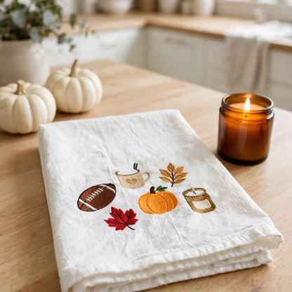 A folded white linen kitchen towel with autumn-themed embroidery sits on a wooden table in a warm kitchen setting. The embroidery features a football, a mug, a pumpkin, a leaf, a candle jar, and a red maple leaf.