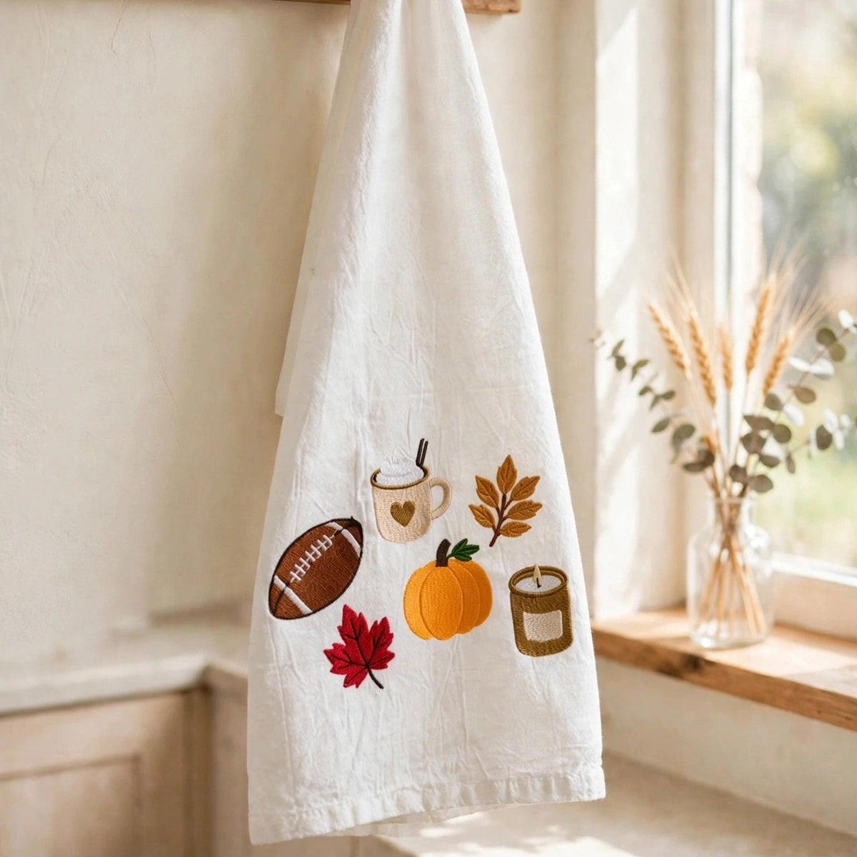 A white kitchen towel with autumn-themed embroidered motifs including a football, mug, pumpkin, leaves, and candle, hanging from a wooden peg rack next to a window.