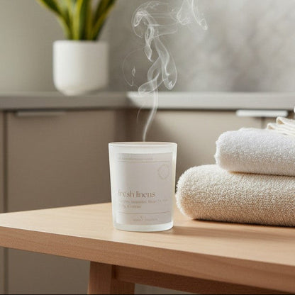 A frosted glass candle jar with visible steam rising from it sits on a light wood table next to a stack of folded cream towels in a softly lit bathroom or spa setting.