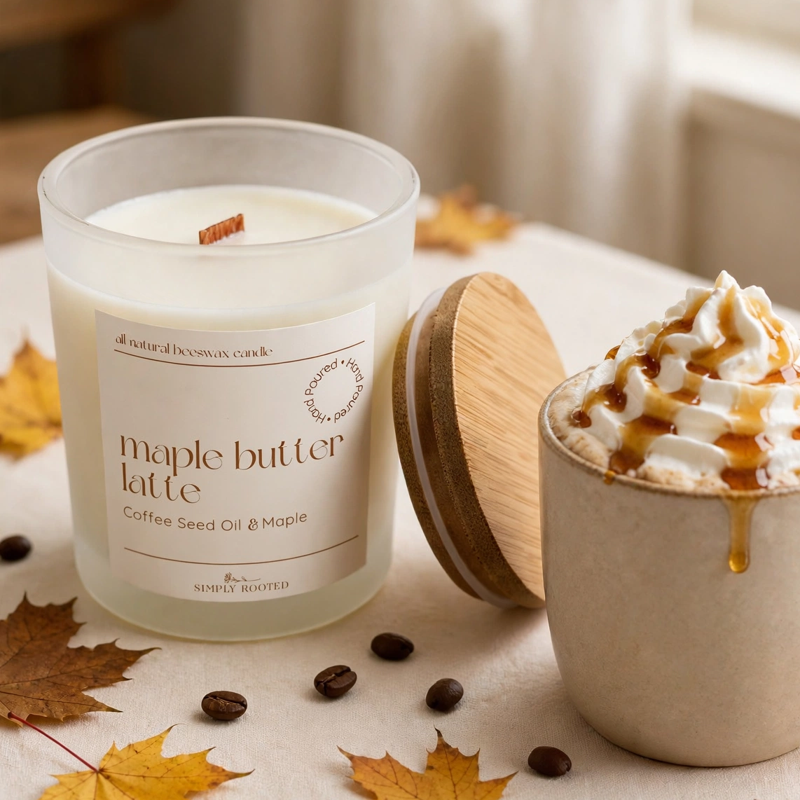 A candle jar labeled maple butter latte sits beside a ceramic mug filled with a whipped latte topped with caramel drizzle, on a table with autumn leaves and coffee beans.