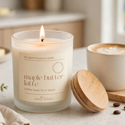 A lit glass jar candle labeled 'maple butter latte' sits on a countertop beside a wooden lid, with a cup of latte and croissants blurred in the background.